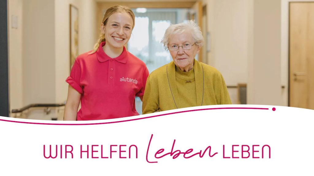 Eine junge Pflegerin steht freundlich lächelnd neben einer älteren Dame mit grauen Haaren und senfgelber Strickjacke - „Wir helfen Leben leben“.