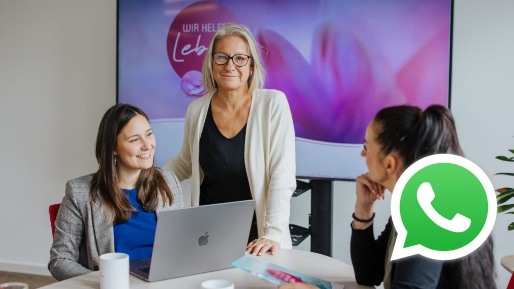 Drei Mitarbeiterinnen von aiutanda Süd-Ost im Gespräch am Laptop – im Vordergrund das WhatsApp-Logo als Hinweis auf den neuen Kommunikationskanal.